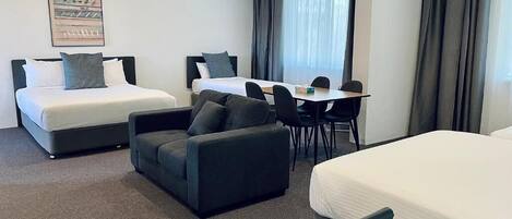 Een hotelkamer met twee bedden, een bank, een eettafel met stoelen en een groot raam met gordijnen.