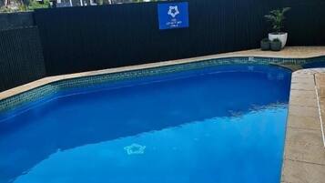 Piscina com cerca escura, placa com logotipo e vista de edifícios ao fundo.
