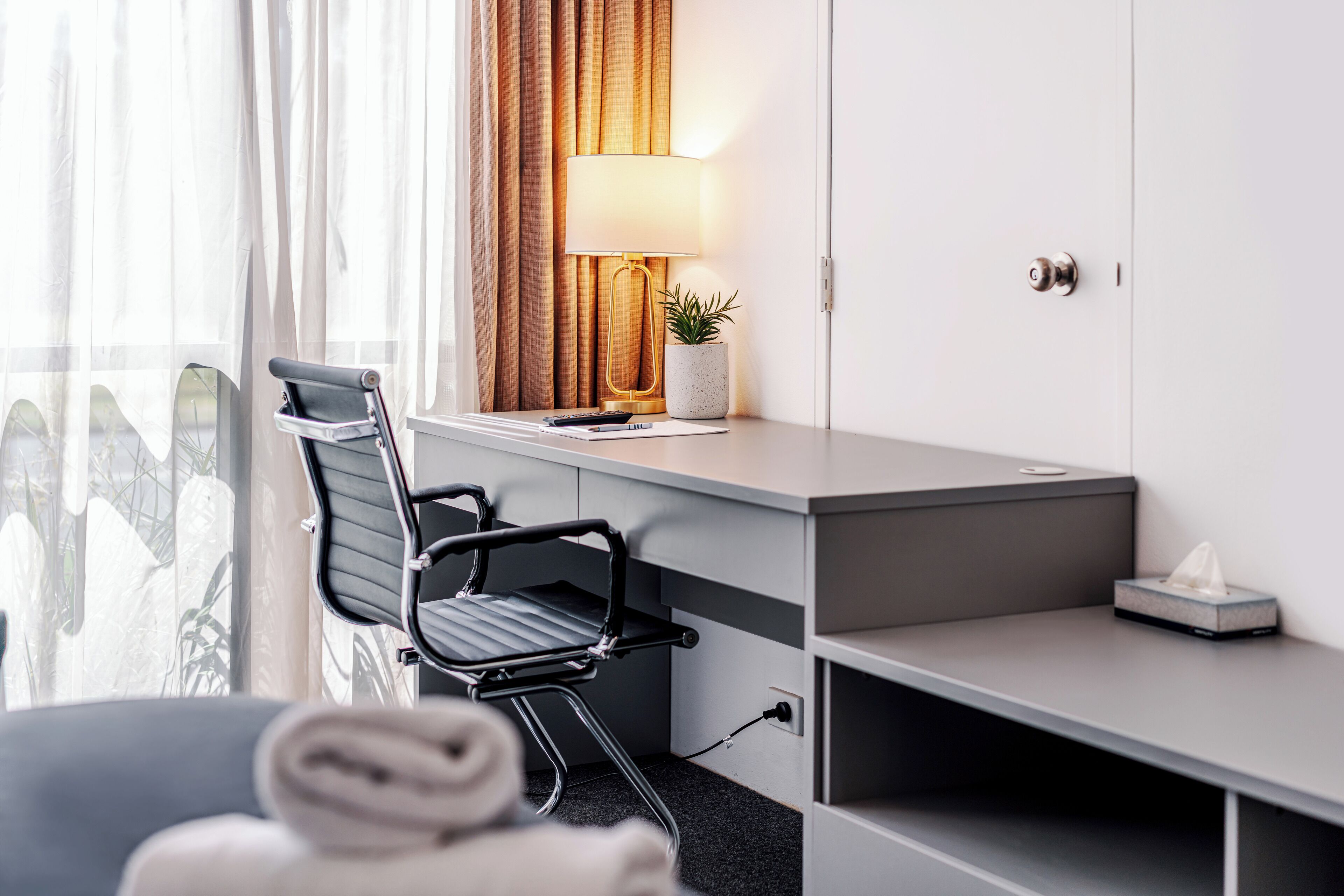A hotel room with a desk, chair, lamp, potted plant, and a view of a cityscape through the window.