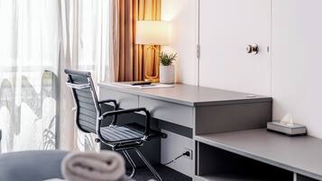 A hotel room with a desk, chair, lamp, potted plant, and a view of a cityscape through the window.