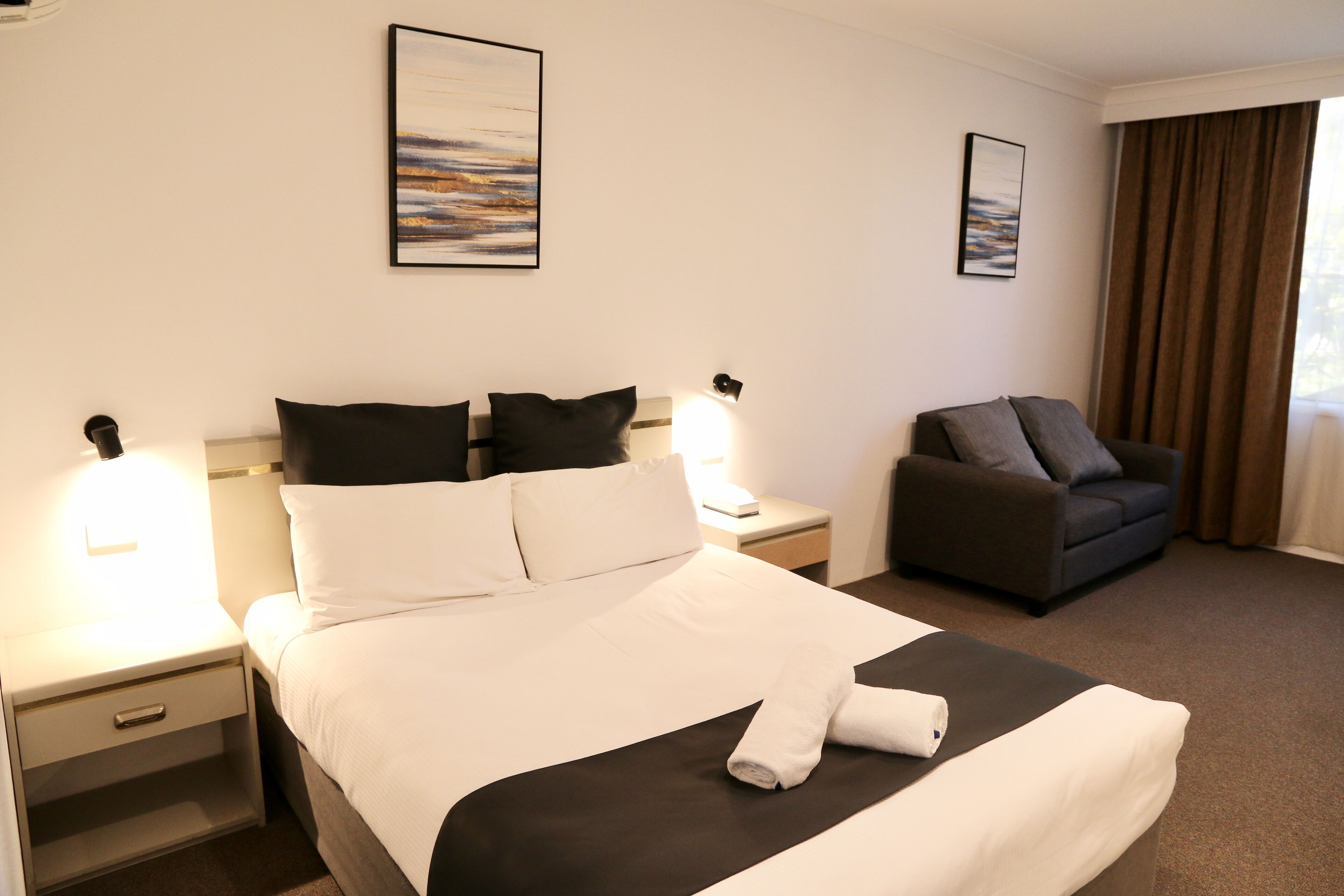 A hotel room with a neatly made bed, a sofa, a nightstand with a lamp, and two framed pictures on the wall.