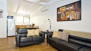 Una sala de estar con dos sofás de cuero negro, piso de madera, una cocina al fondo y un cuadro abstracto grande en la pared.