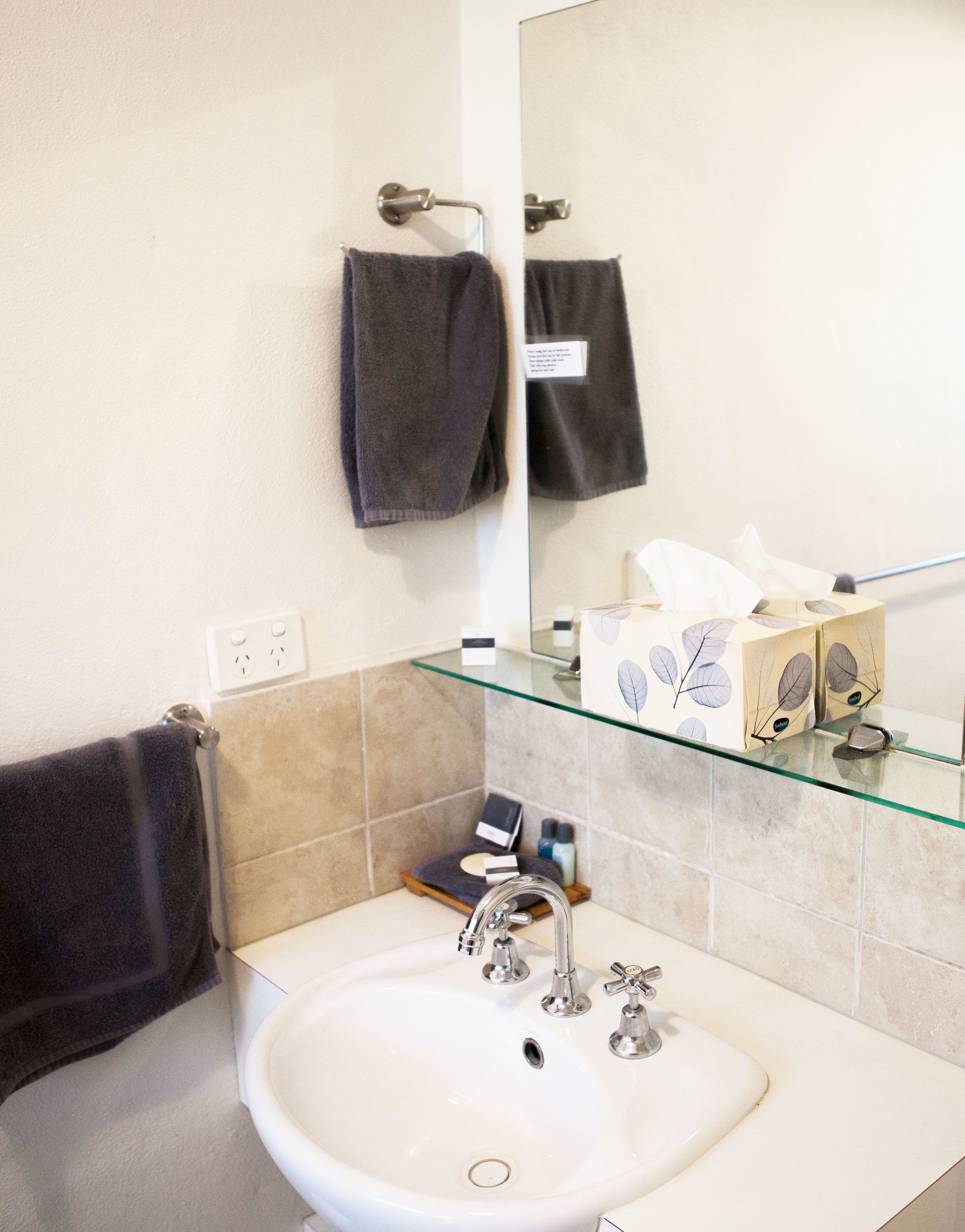 Une salle de bain avec un lavabo blanc, une étagère en verre où sont rangés les articles de toilette, et deux serviettes suspendues à des crochets.