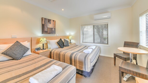 Habitación de hotel con dos camas, una mesita pequeña y una silla. Hay una ventana con persianas, un aire acondicionado de pared y un cuadro en la pared.