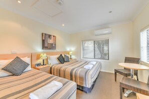 Habitación de hotel con dos camas, una mesita pequeña y una silla. Hay una ventana con persianas, un aire acondicionado de pared y un cuadro en la pared.