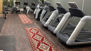 Uma academia com esteiras alinhadas contra uma parede cinza, um tapete com padrão vermelho e equipamentos de ginástica ao fundo.