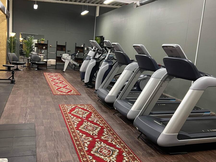 Un gimnasio con cintas de correr alineadas contra una pared gris, alfombra roja estampada y equipo de ejercicio al fondo.