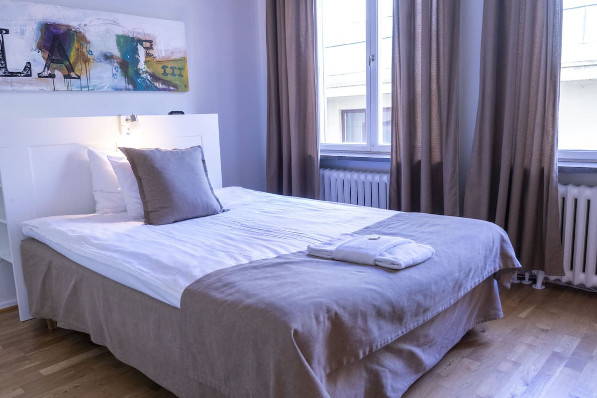 Ein ordentlich bezogenes Bett mit weißer und beiger Bettdecke, ein gefaltetes Handtuch und zwei Kissen. Darüber am Bett hängt ein großes abstraktes Gemälde.