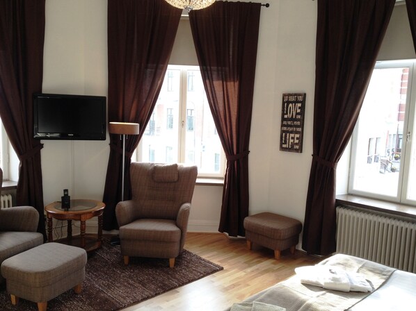 A living room with a brown armchair, a small round table, a television, and a window with curtains. - Stadshotellet Kristianstad (Kristianstad)