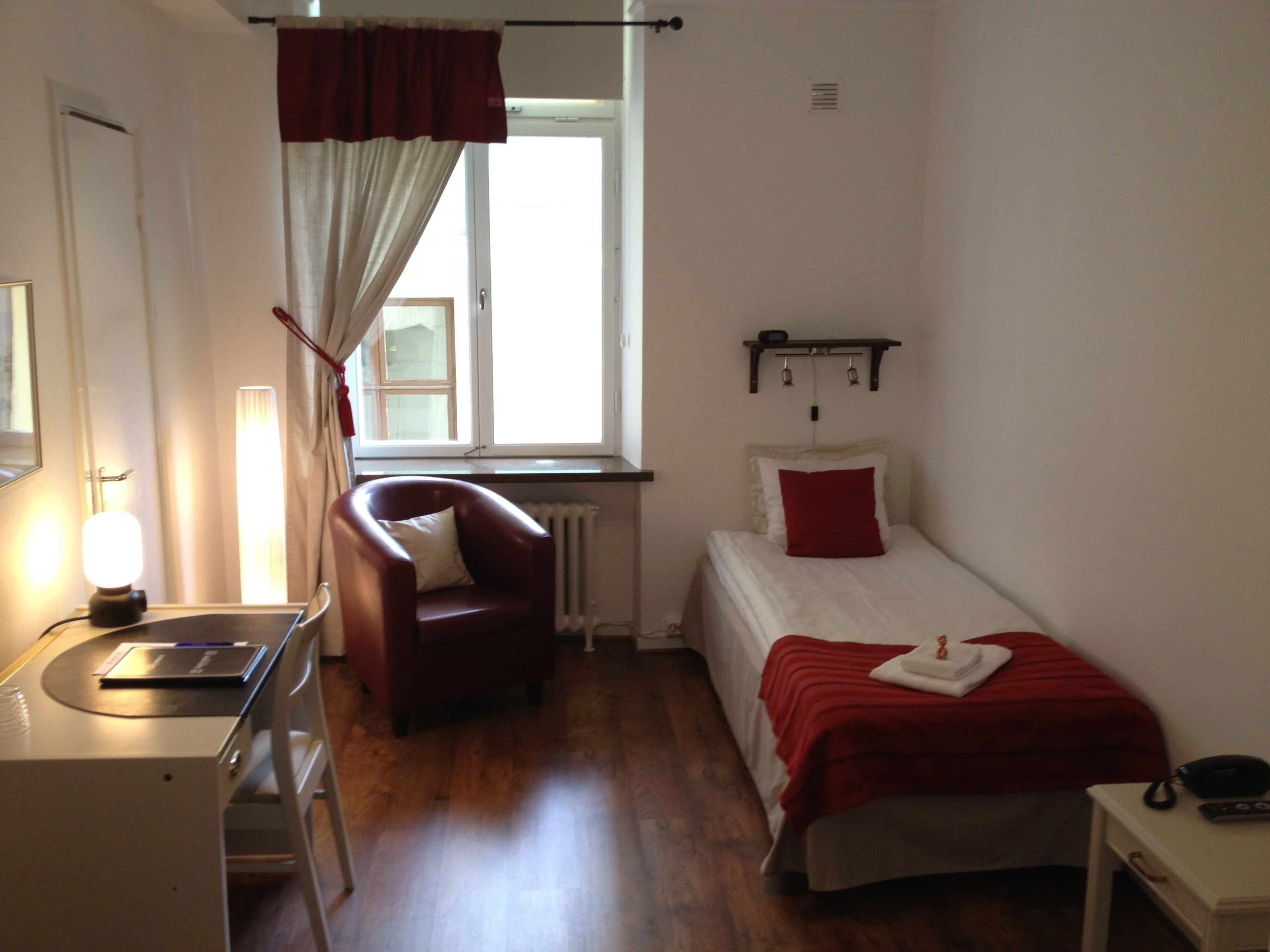 A small, well-lit room with a single bed, a chair, a desk with a lamp, and a window with curtains.