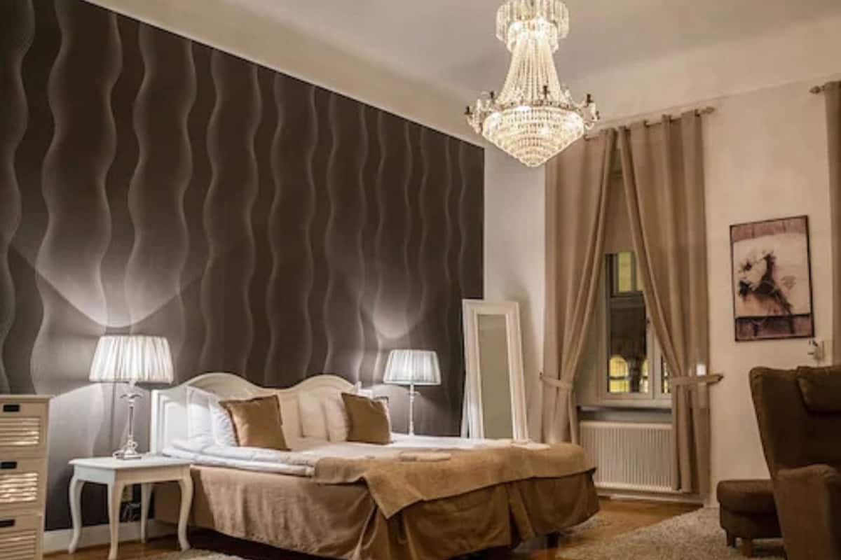 Ein Schlafzimmer mit einem Bett, zwei Nachttischen, einer Kristallleuchter, einem Fenster mit Vorhängen und einer Heizung.