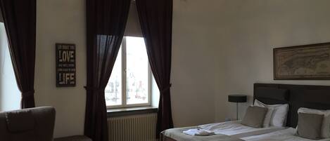 Un dormitorio con una cama, una silla, un radiador, una ventana con cortinas y un candelabro.