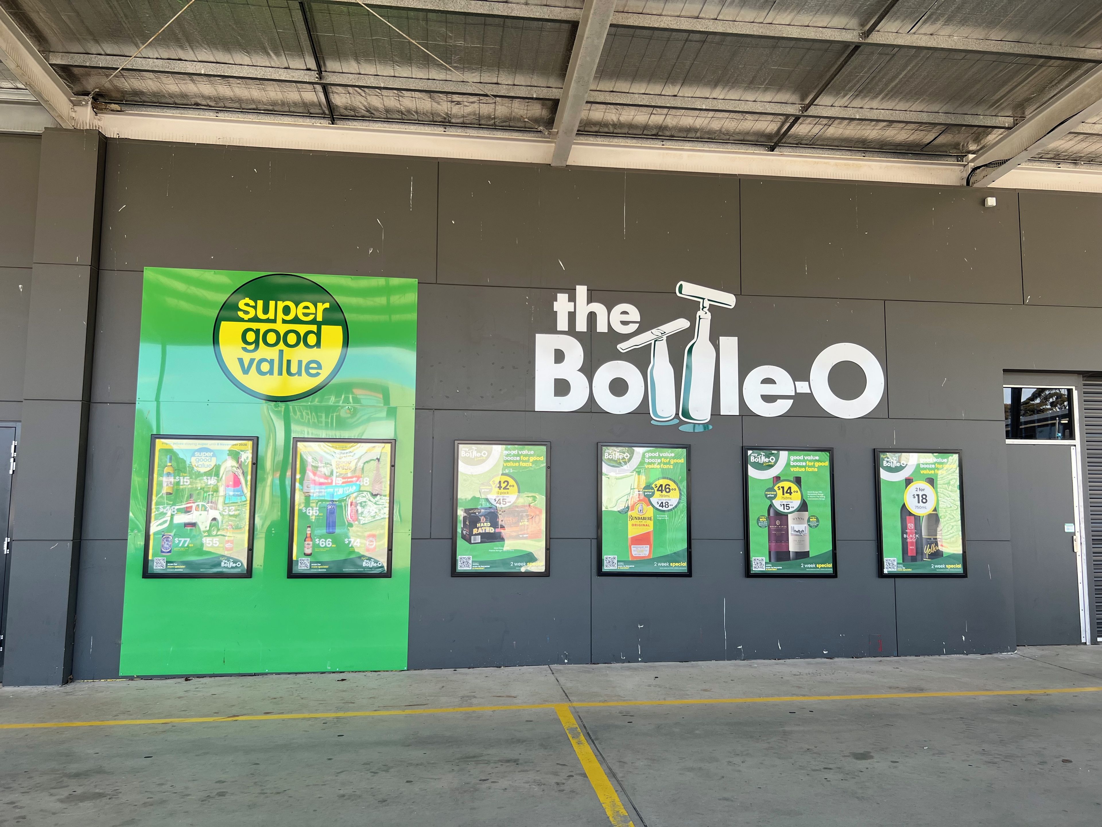 A wall with advertisements for 'The Bottle-O' and 'super good value' promotions, featuring price specials and product images.