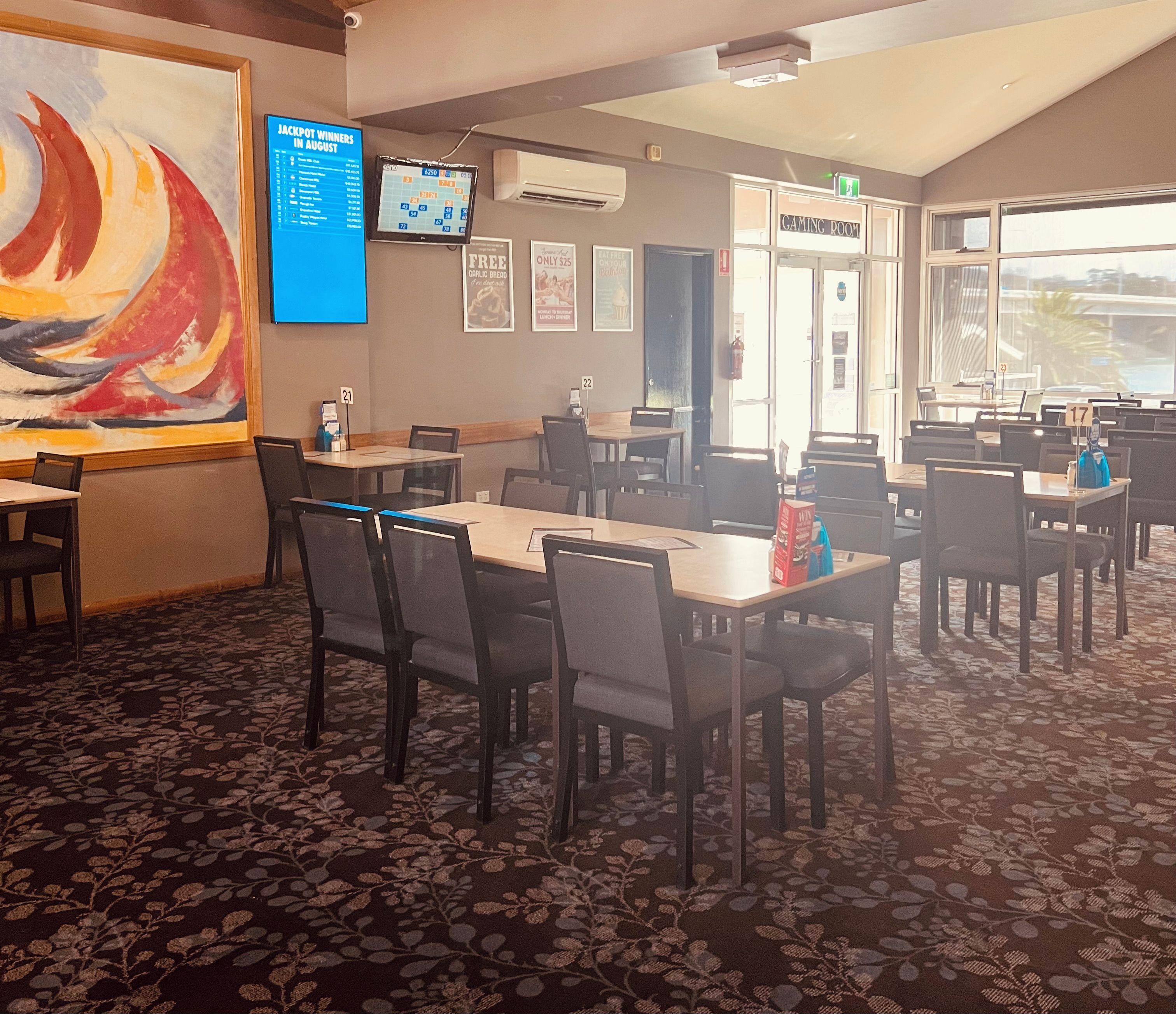 Un restaurante con mesas y sillas, un cuadro grande en la pared, una pantalla mostrando información, y una puerta con la leyenda 'GAMING ROOM'.