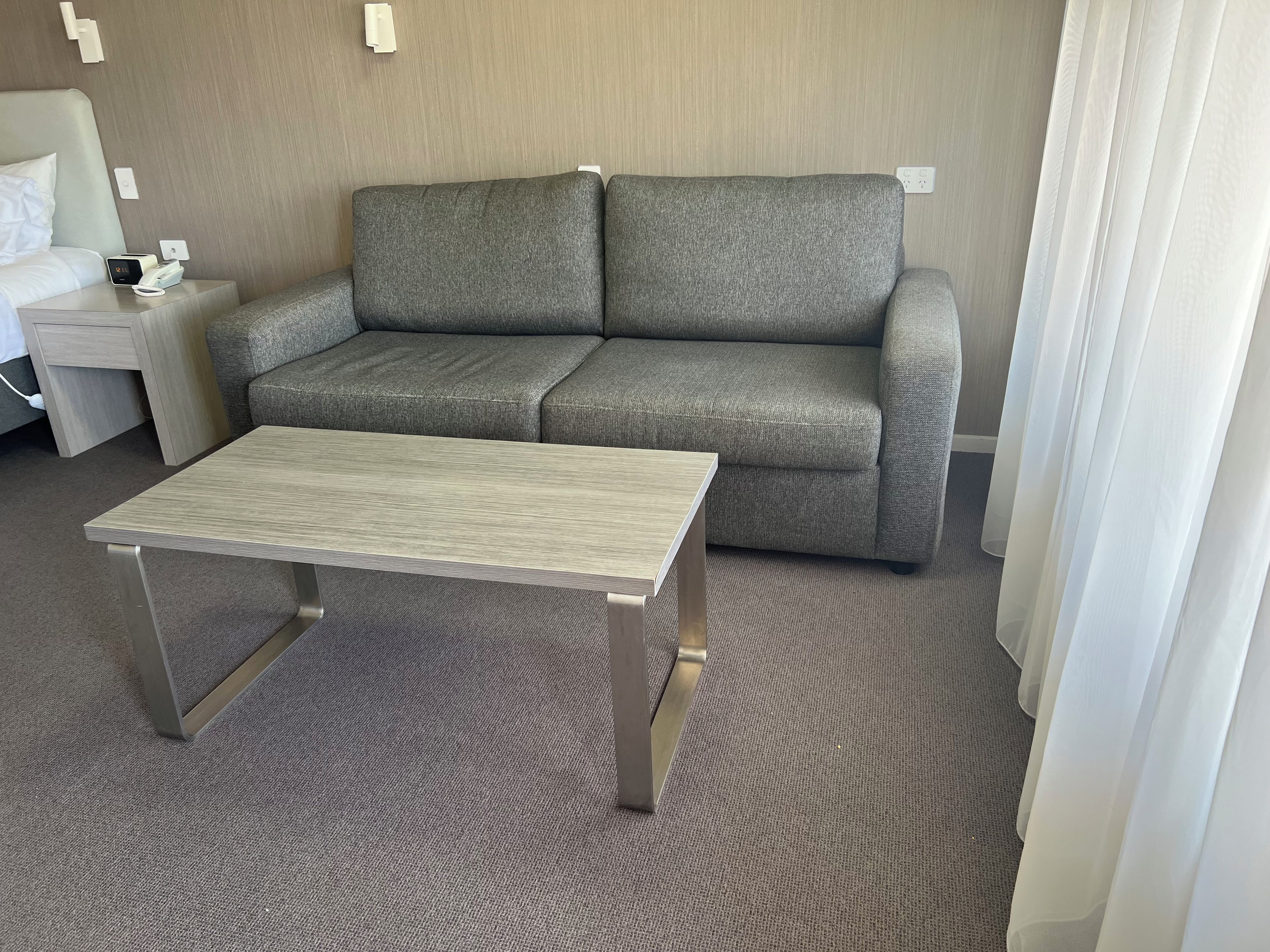 Una habitación de hotel con un sofá gris, una mesita, una mesita de noche con una lámpara y un enchufe en la pared.