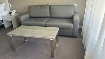 Una habitación de hotel con un sofá gris, una mesita, una mesita de noche con una lámpara y un enchufe en la pared.