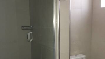 Un baño con ducha separada por mampara de vidrio, inodoro blanco y un estante pequeño.