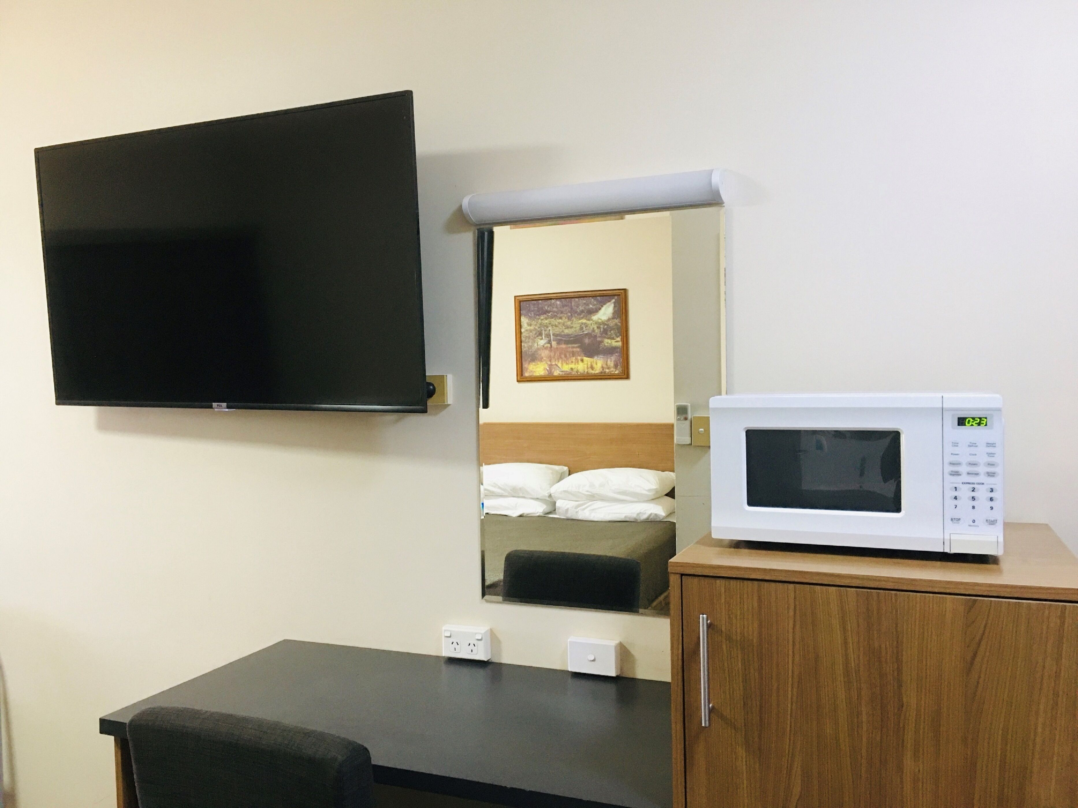 A hotel room with a flat-screen TV mounted on the wall, a microwave on a wooden cabinet, a bed with white bedding, and a painting on the wall.