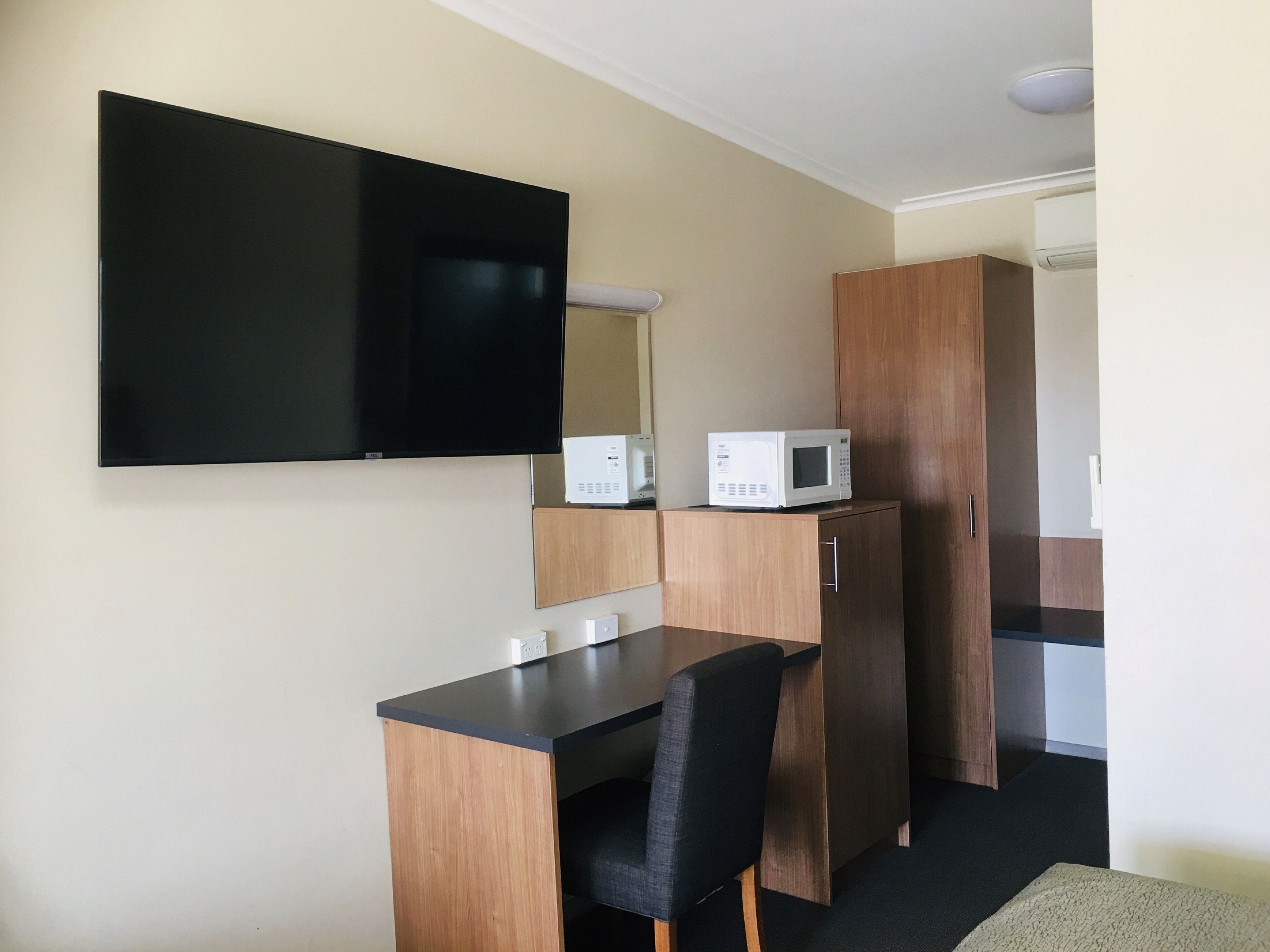Una habitación pequeña y moderna con una televisión de pantalla plana montada en la pared, un microondas en un mueble y un escritorio con una silla.