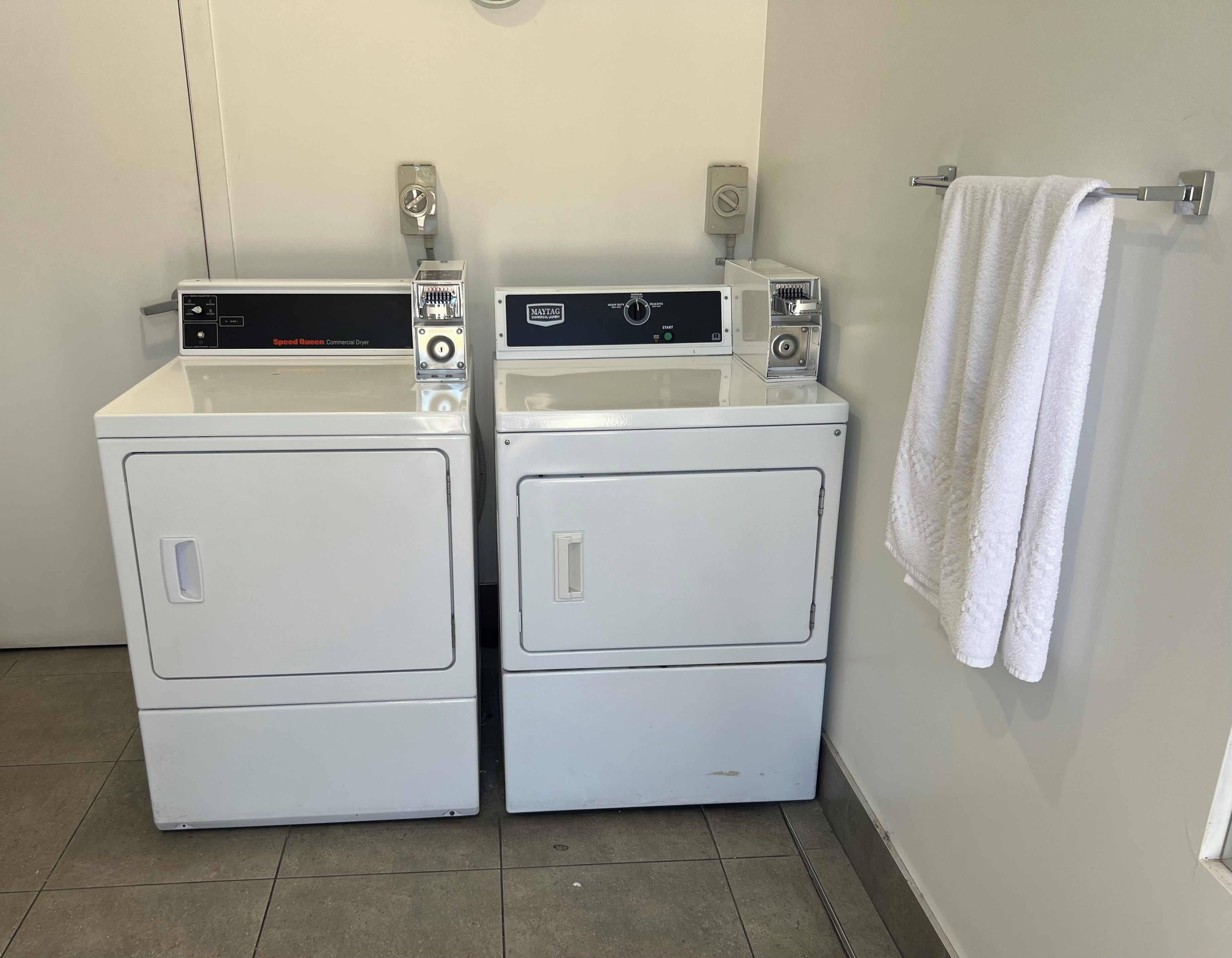 Duas secadoras comerciais brancas com painéis de controle, uma toalha branca pendurada em um suporte e um piso revestido de azulejos.