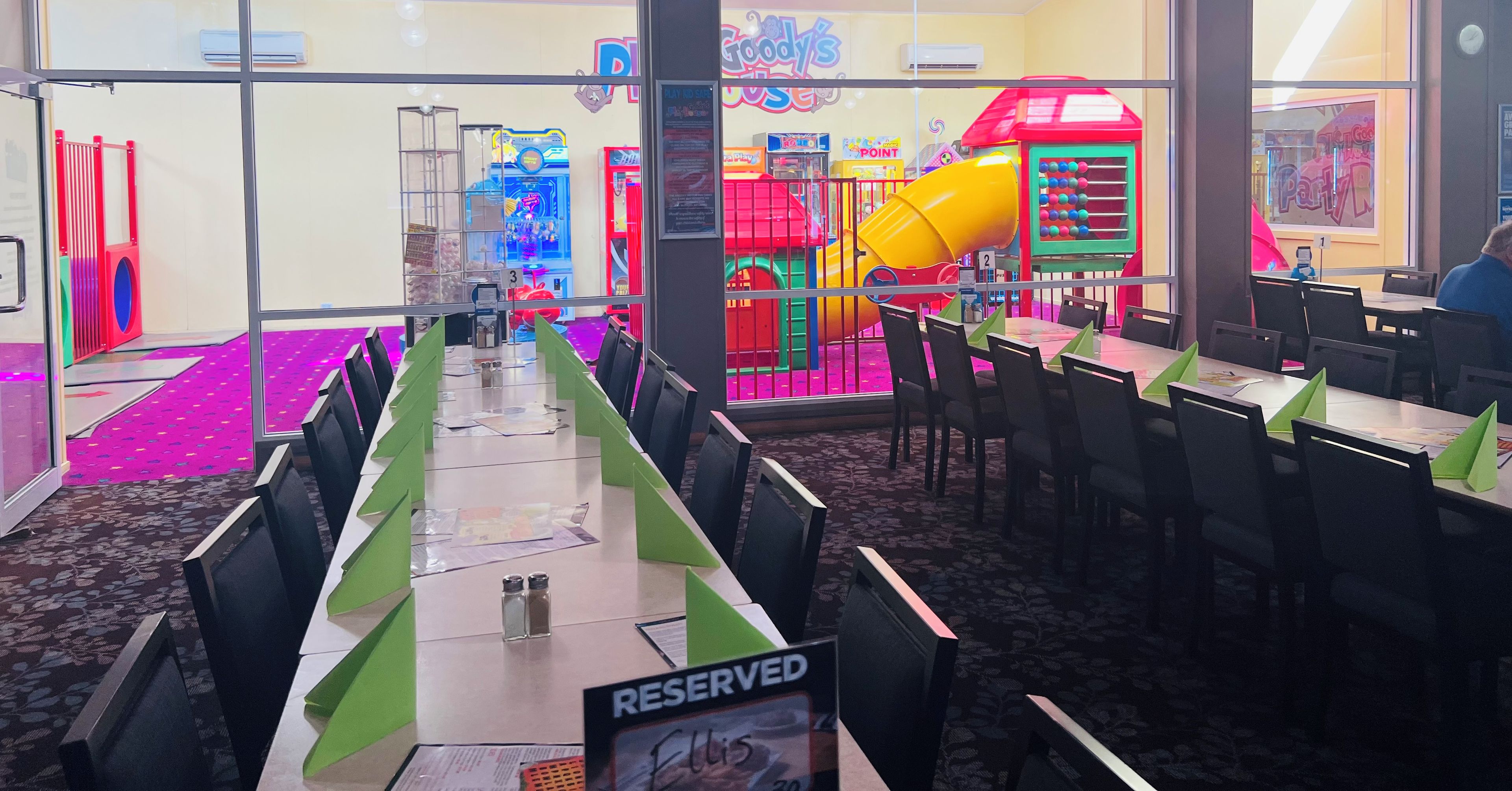 Un restaurante con vista a un área de juegos interior. Las mesas están dispuestas con posavasos verdes y se ve un cartel de 'RESERVED'.