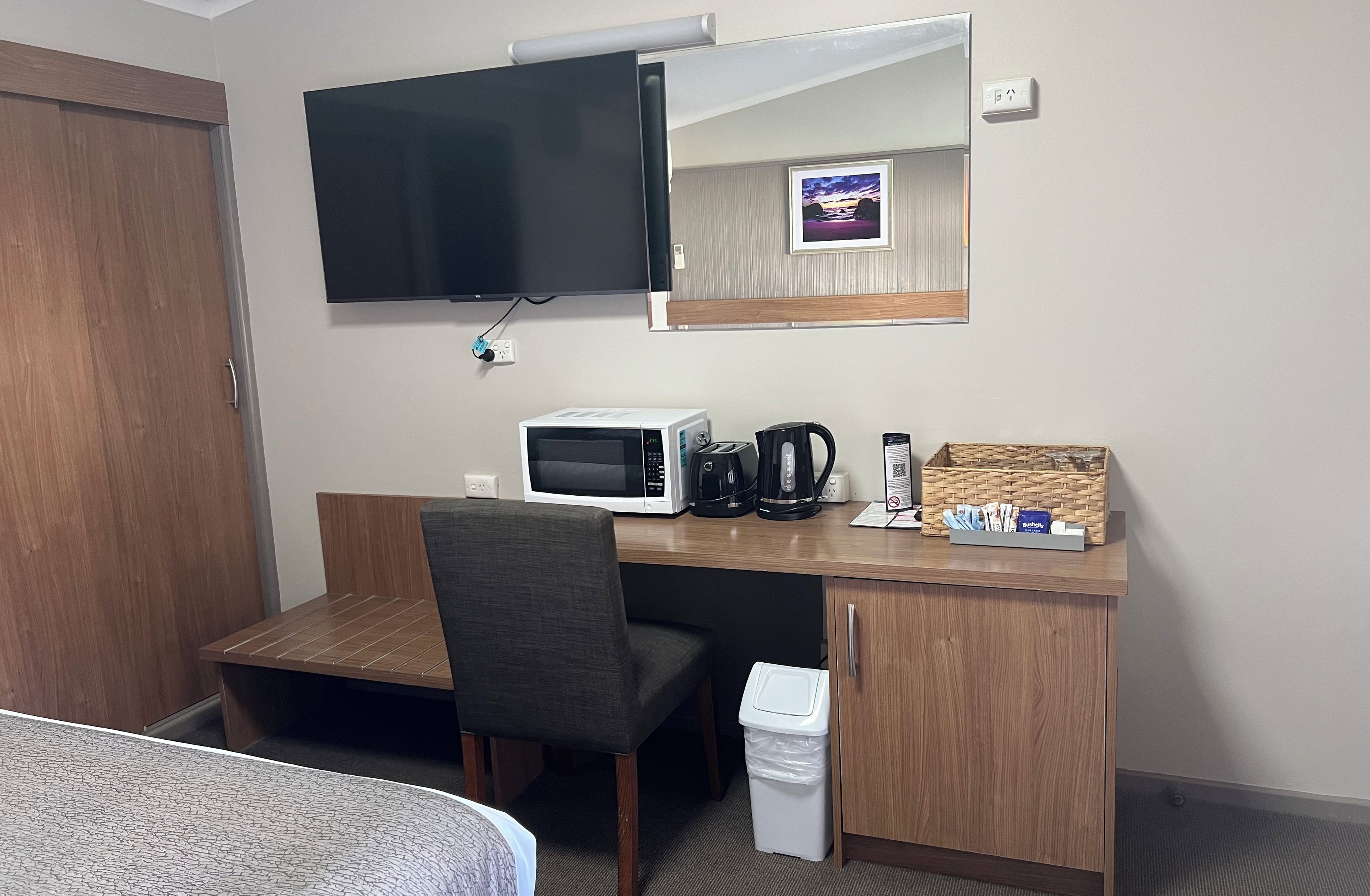A hotel room with a bed, a desk with a microwave, a coffee maker, a trash bin, and a television mounted on the wall.