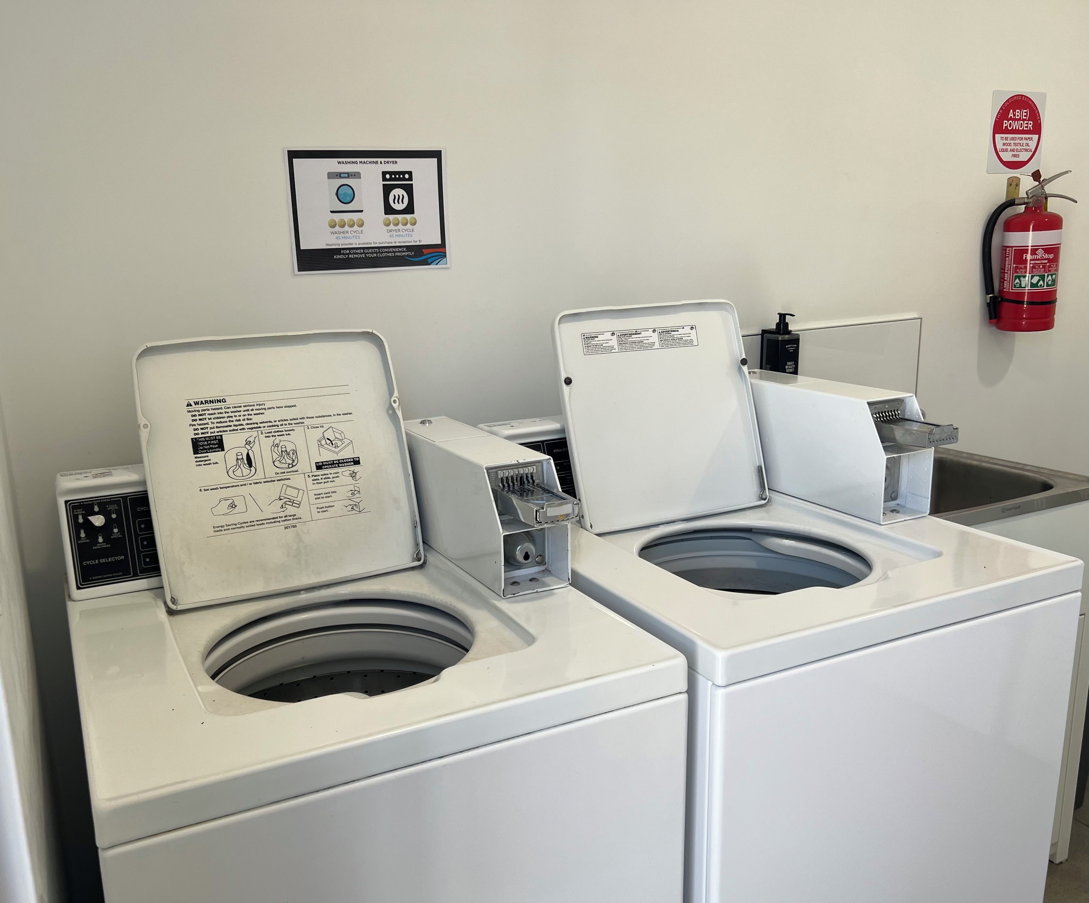 Une laverie publique équipée de deux machines à laver et d’un sèche-linge, d’un extincteur fixé au mur et d’un panneau indiquant l’utilisation d’un extincteur à poudre de type A:B(E).