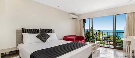 A modern hotel room with a large bed, a red chair, a desk, and a balcony with a view of the ocean.