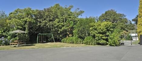 A parking area with a swing set and a bench, surrounded by dense greenery and trees.