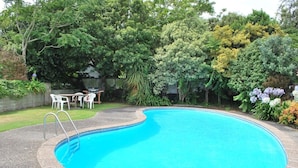 En swimmingpool med en buet kant, omgivet af en belagt terrasse. Hvide plaststole og et bord ses i baggrunden.