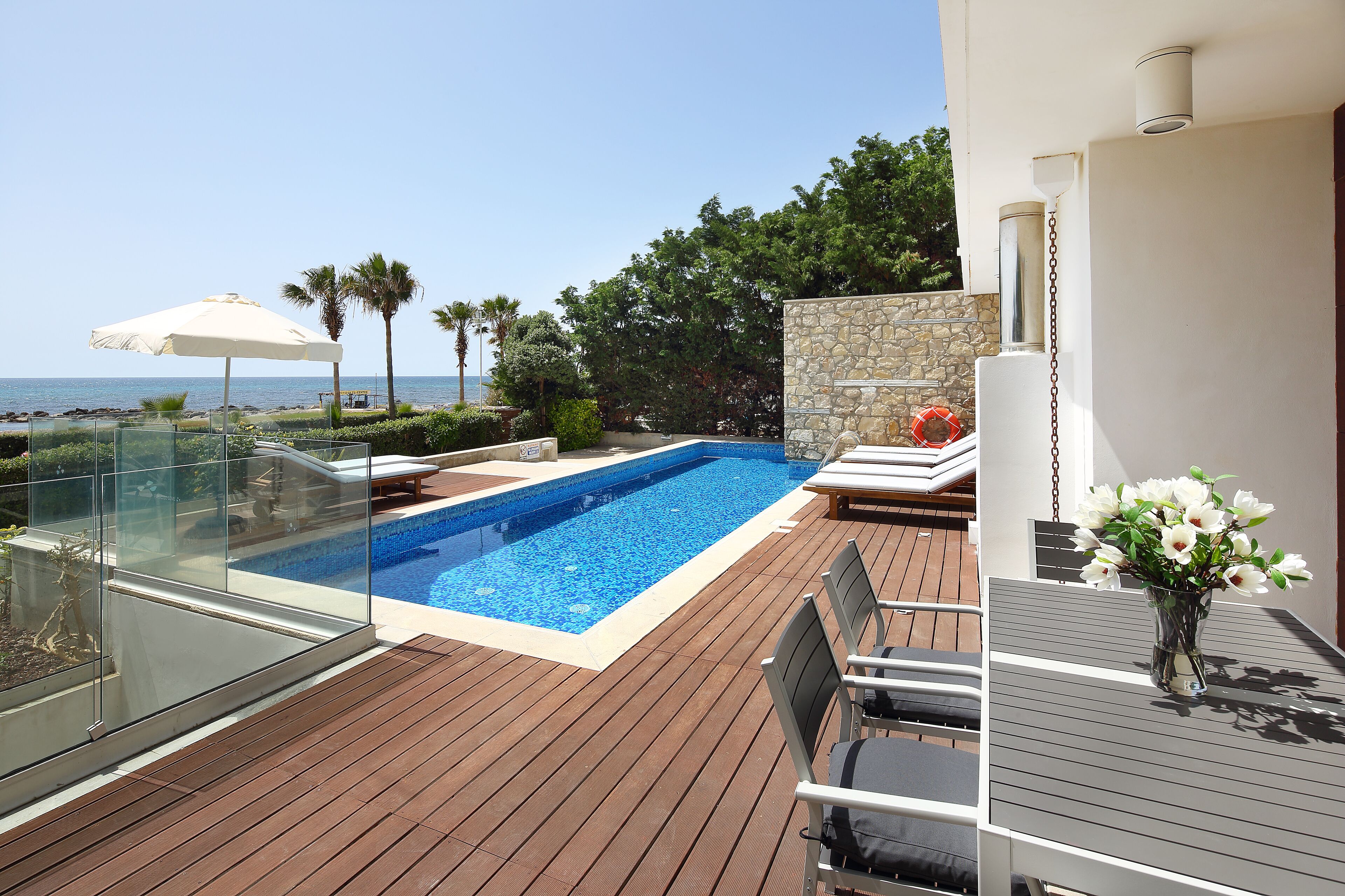 A rooftop pool area with a glass balustrade, lounge chairs, and a table with a vase of flowers.