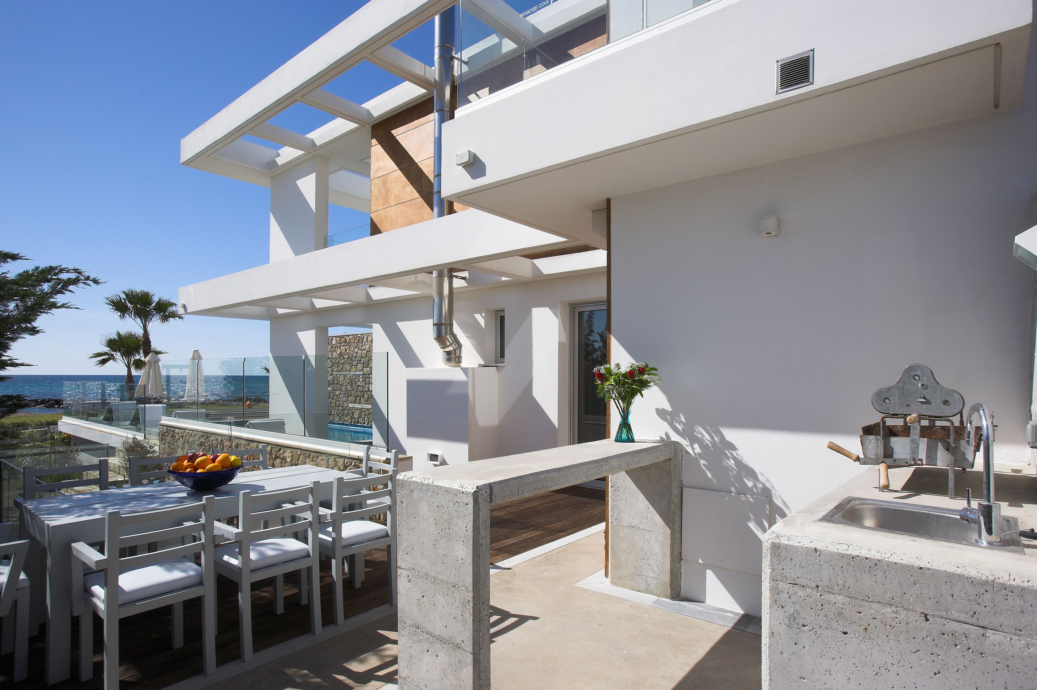 A modern outdoor dining area with a concrete table and chairs, a bowl of fruit, and a view of the ocean.
