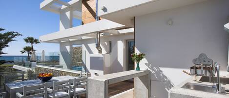 A modern outdoor dining area with a concrete table and chairs, a bowl of fruit, and a view of the ocean.