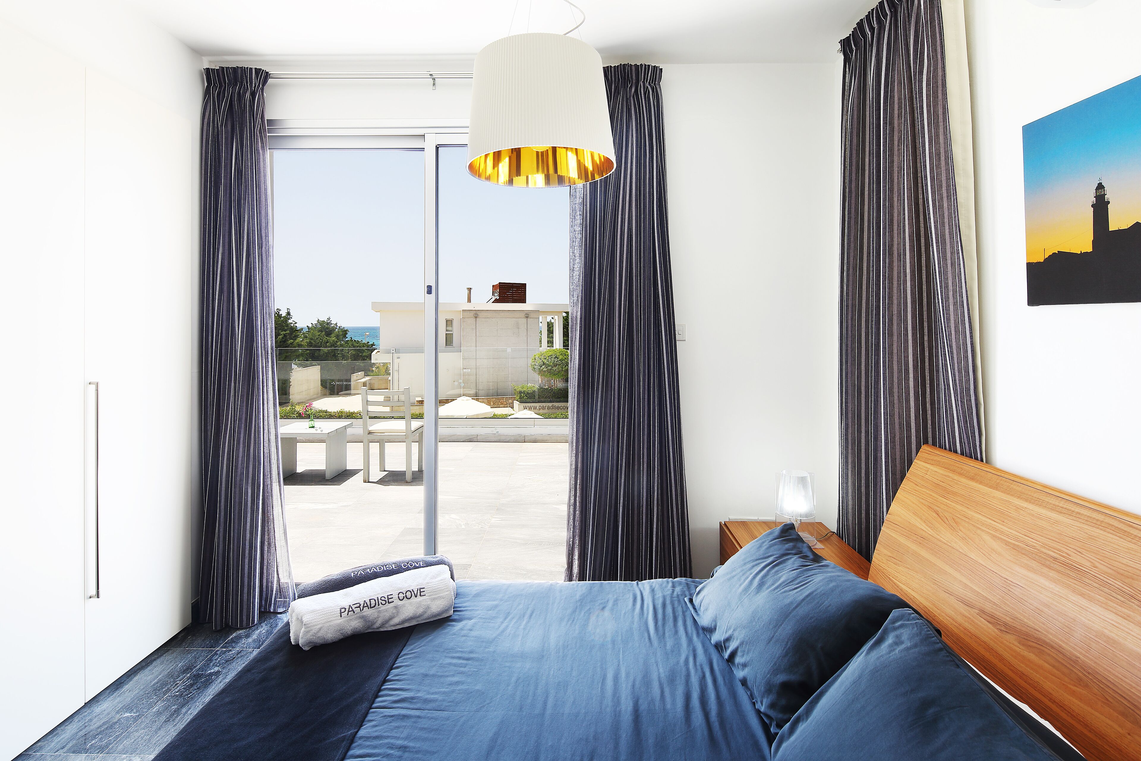 Un dormitorio con cama, mesita de noche, lámpara y vista a un patio exterior.