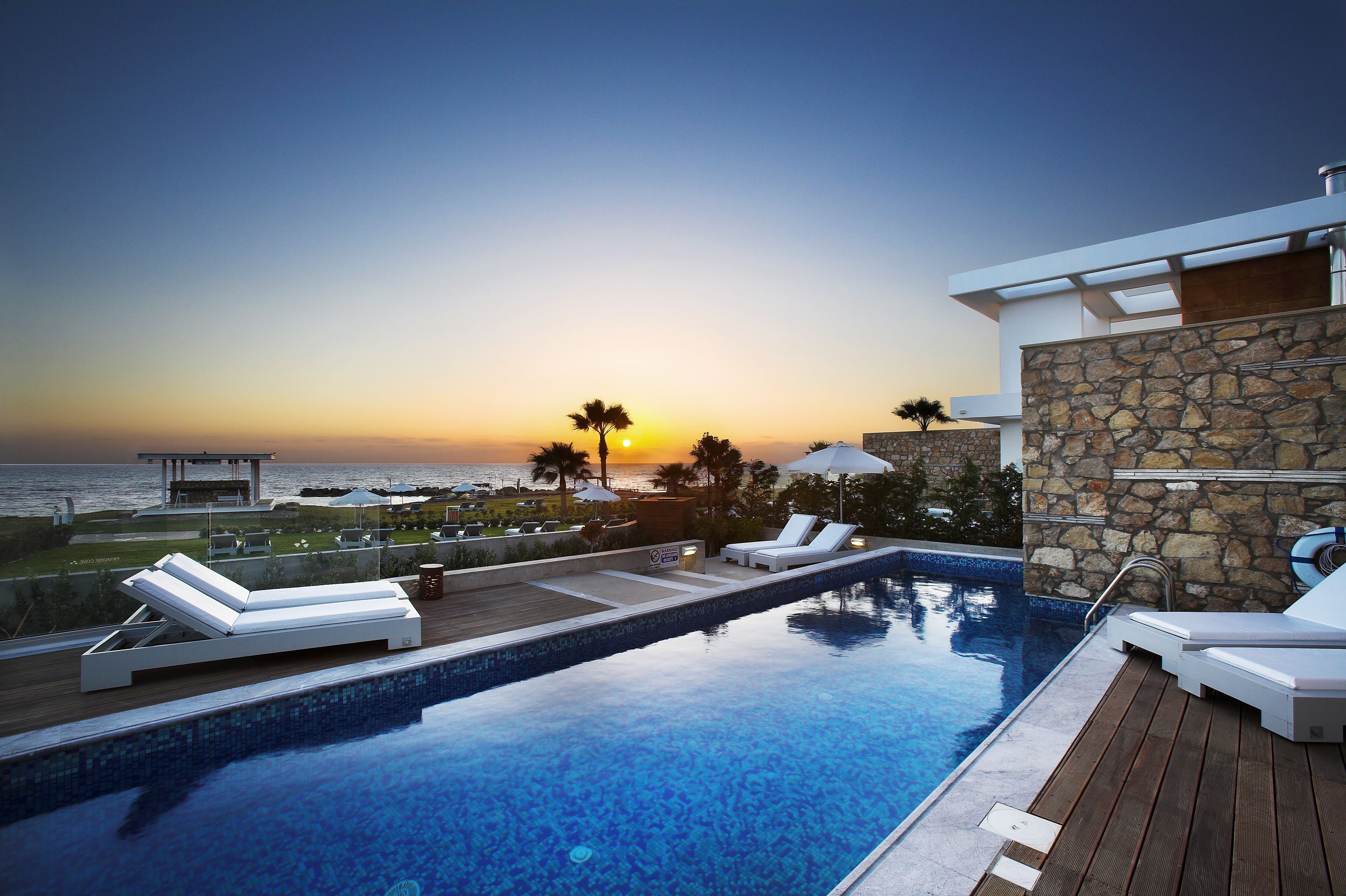 Un área de piscina con sillones de descanso, un muro de piedra y vistas a la puesta de sol sobre el océano.