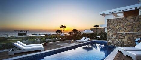 Un área de piscina con sillones de descanso, un muro de piedra y vistas a la puesta de sol sobre el océano.