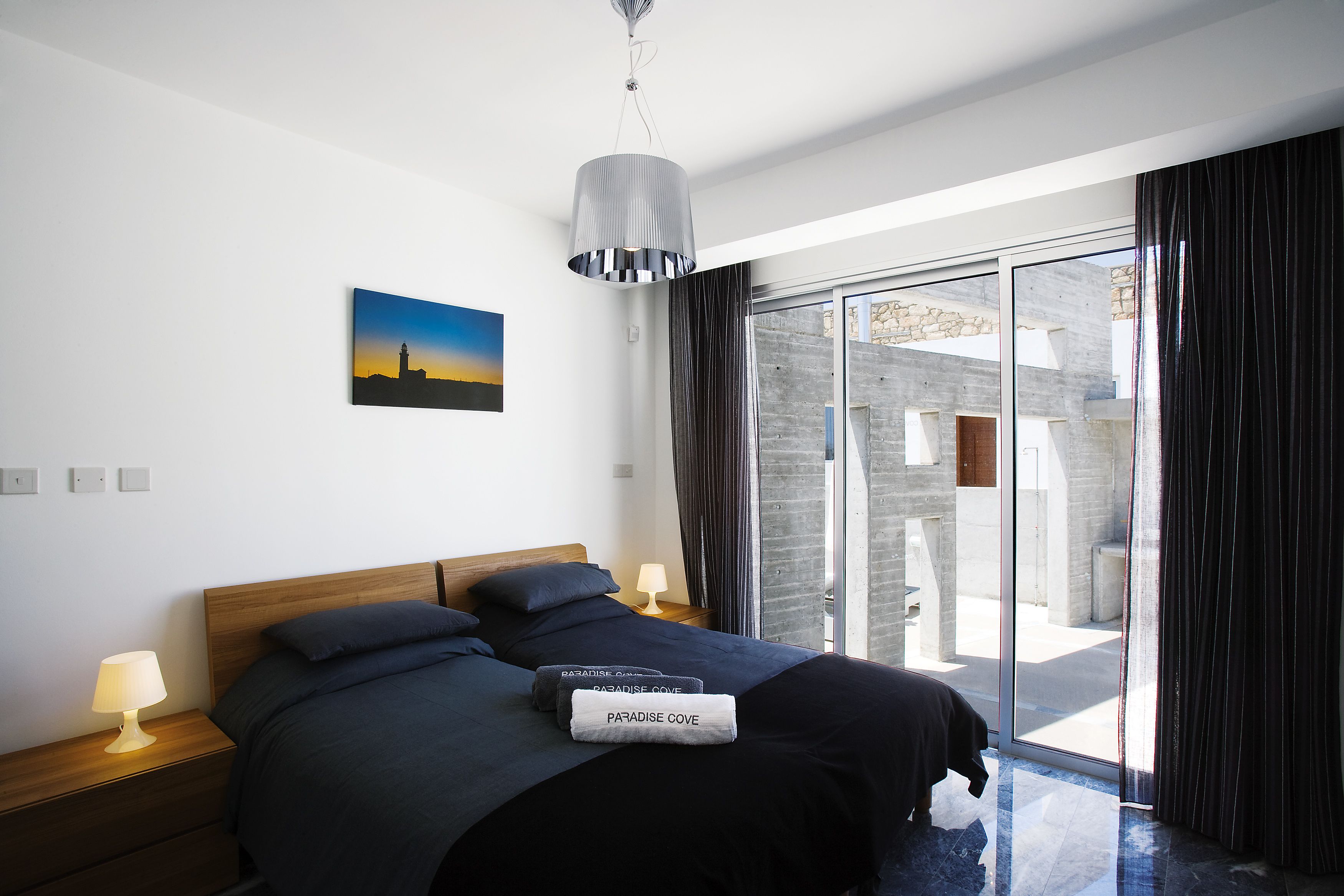 Ein modernes Schlafzimmer mit einem großen Bett, einem Nachttisch mit Lampe, einem Bild an der Wand und Blick auf einen Außenbereich durch eine Glasdoor.