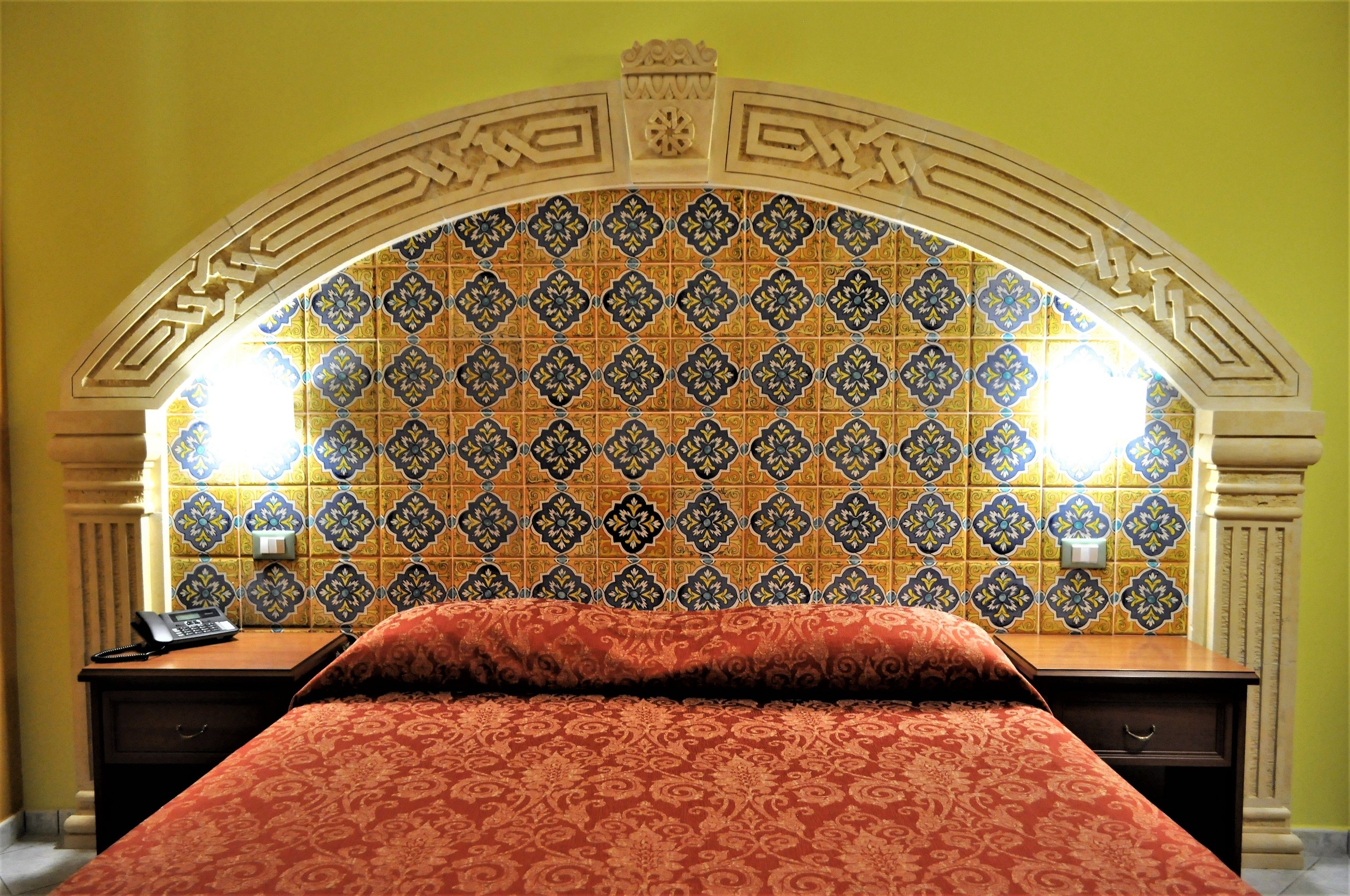 A hotel room with a patterned tile wall, a bed with a red patterned bedspread, a nightstand with a phone, and two wall-mounted lamps.