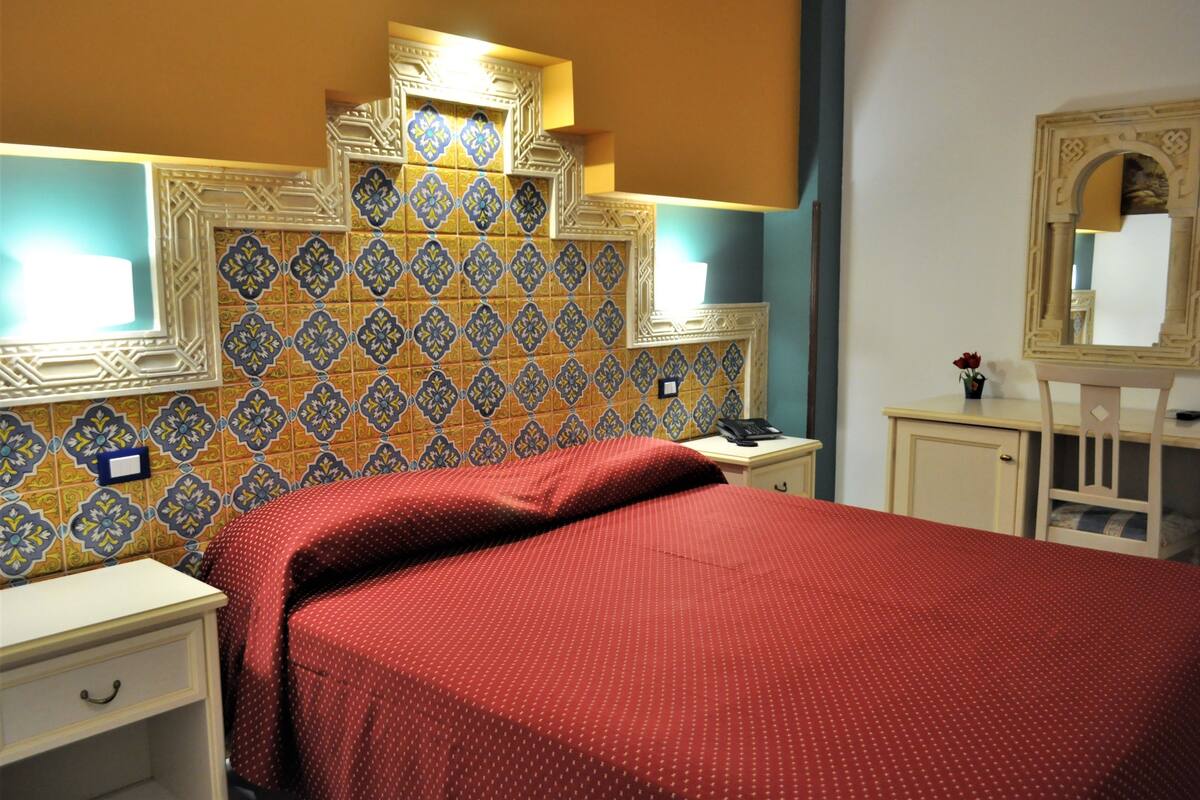 A hotel room with a patterned tile wall, a bed with a red bedspread, a nightstand with a lamp, and a small table with a vase of flowers.