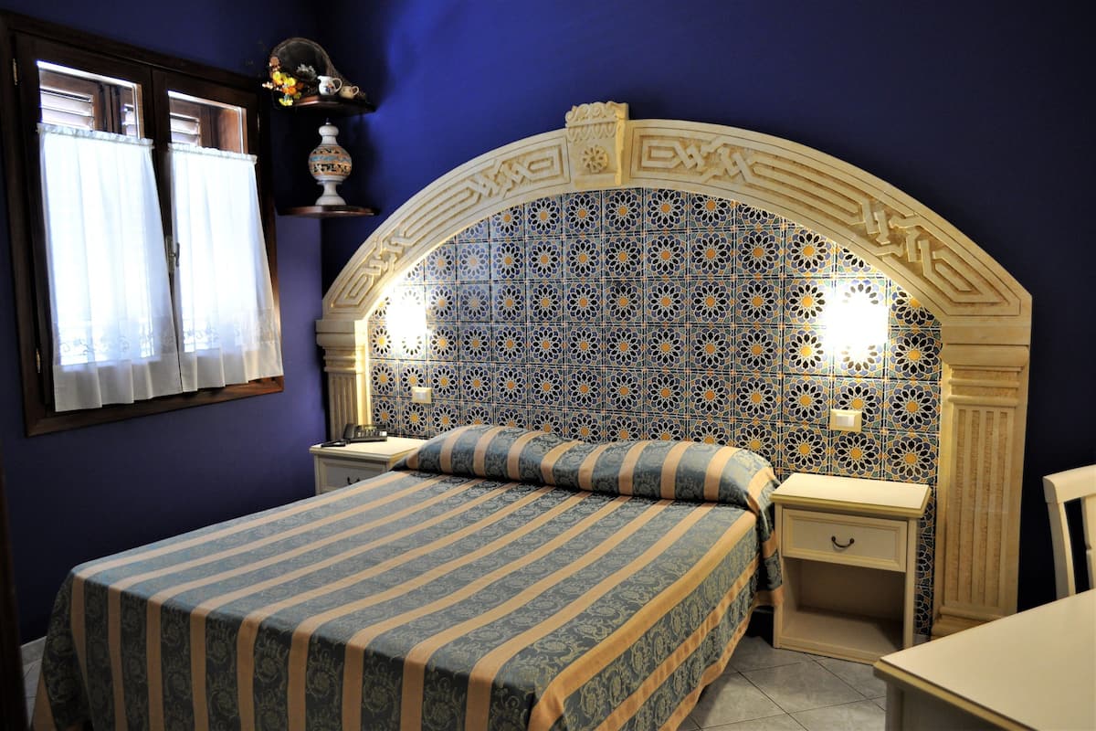 A bedroom with a bed, two bedside tables, a nightstand, a decorative arched ceiling, and a window with curtains.
