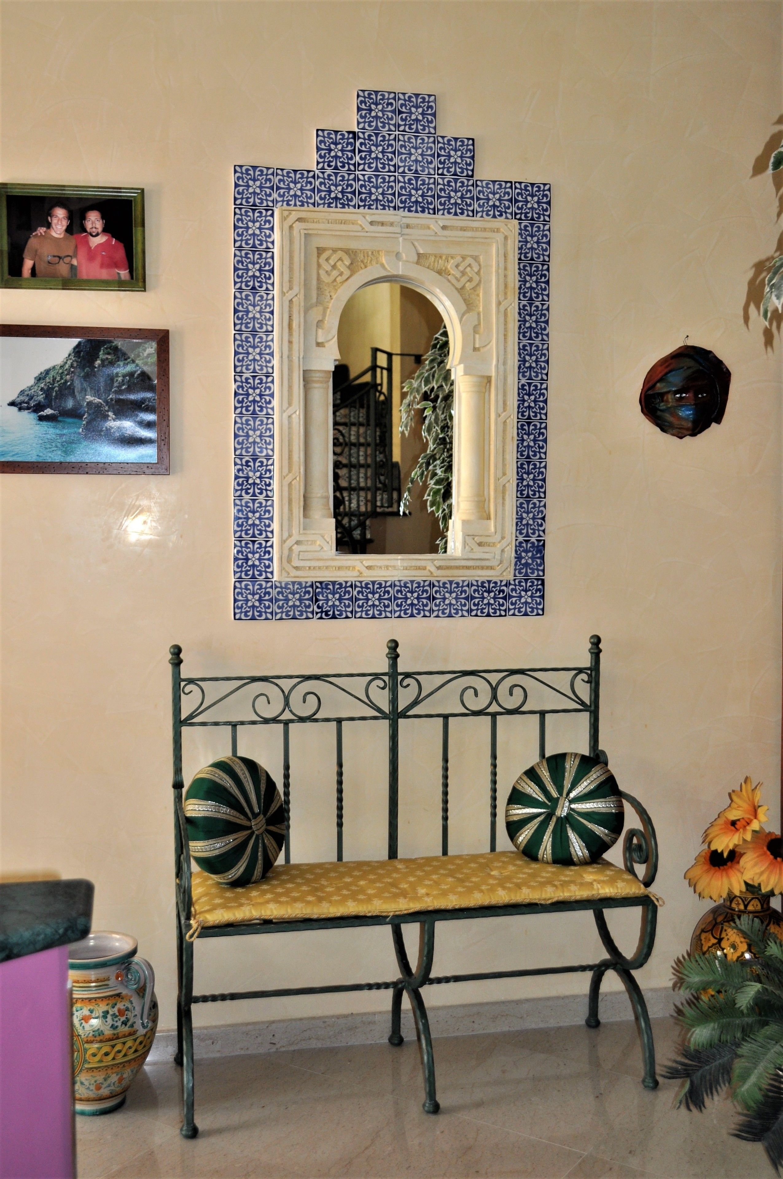 A bench with decorative pillows, a mirror with a blue tile border, and a framed photo on the wall.