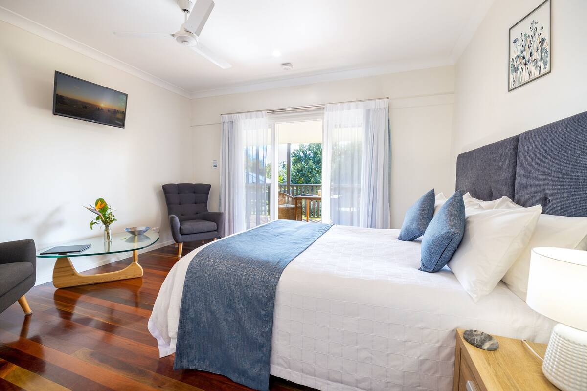 A bedroom with a large bed, a chair, a small table, and a view of the outdoors through a window.