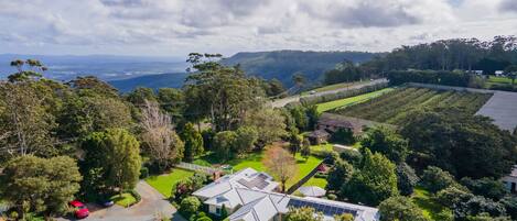 An aerial view of a residential area with a large house featuring solar panels, surrounded by lush greenery and a hilly landscape in the background.