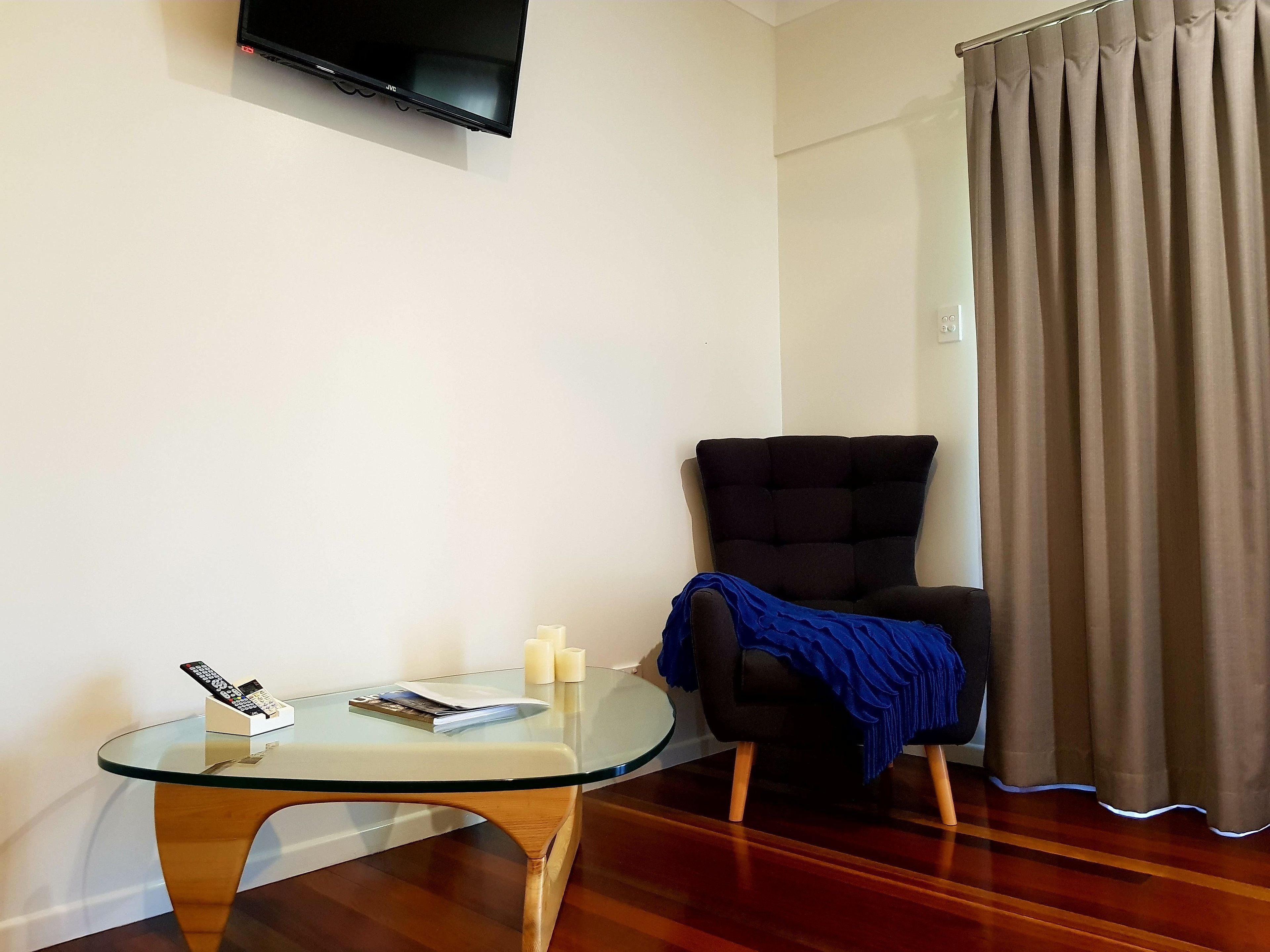 A living room with a glass table, a black armchair, a television mounted on the wall, and a window with curtains.