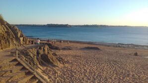 Una playa arenosa con escalones que bajan hacia el agua, cielo despejado y una costa distante con edificios.