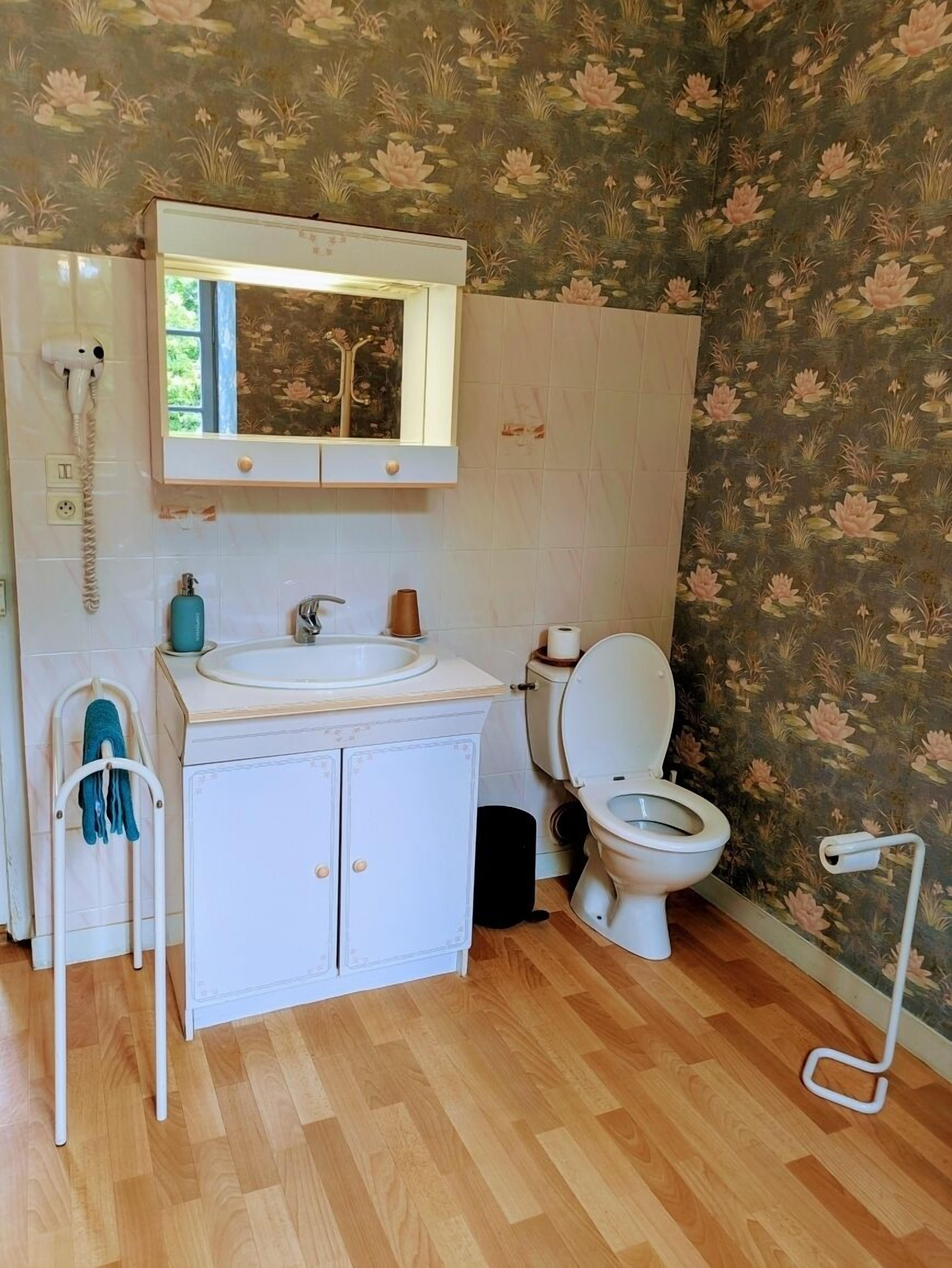 Un baño con lavamanos blanco, espejo, inodoro, suelo de madera y papel tapiz floral.