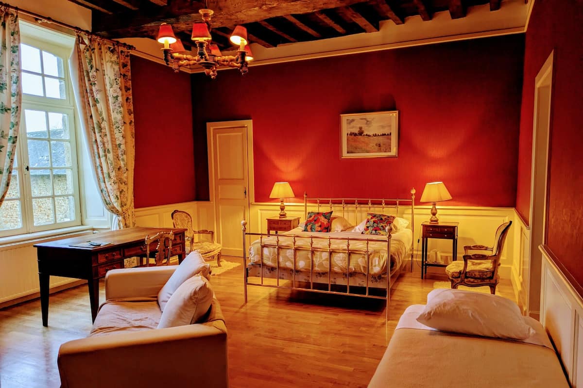 Une pièce avec un plancher en bois, un lit avec un cadre en métal, deux tables de chevet avec des lampes, une chaise, un bureau et une fenêtre avec des rideaux.