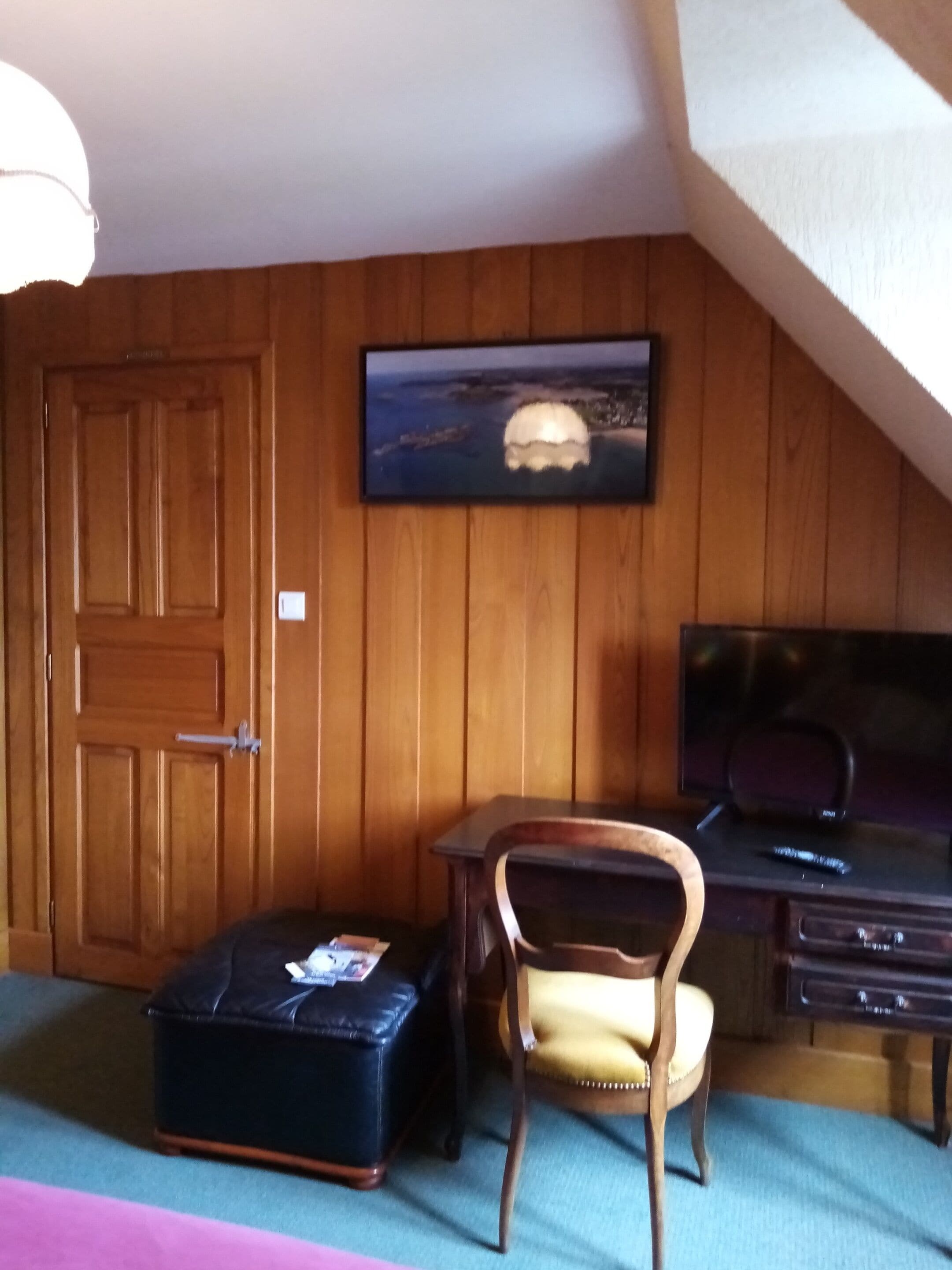 Una habitación con paneles de madera, una puerta, un cuadro enmarcado, un escritorio con una silla, un televisor y un pequeño reposapiés.