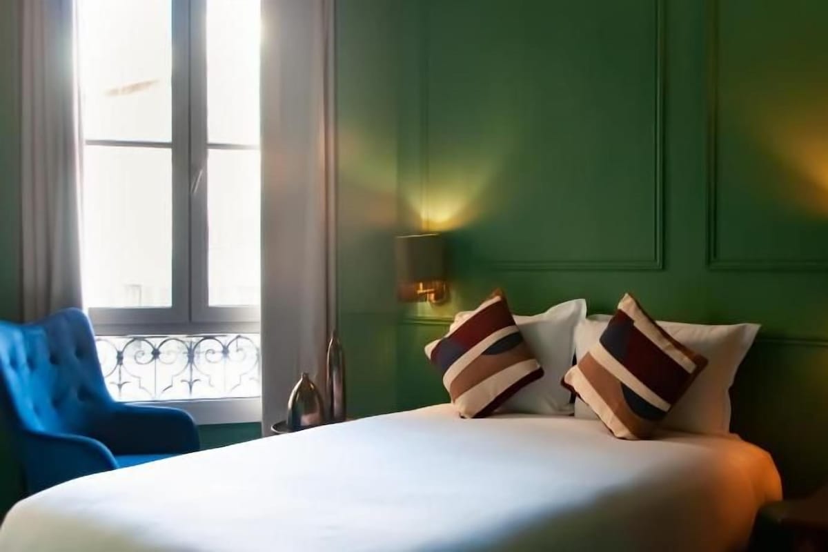 A hotel room with a bed, two bedside lamps, a window with curtains, and a blue chair.