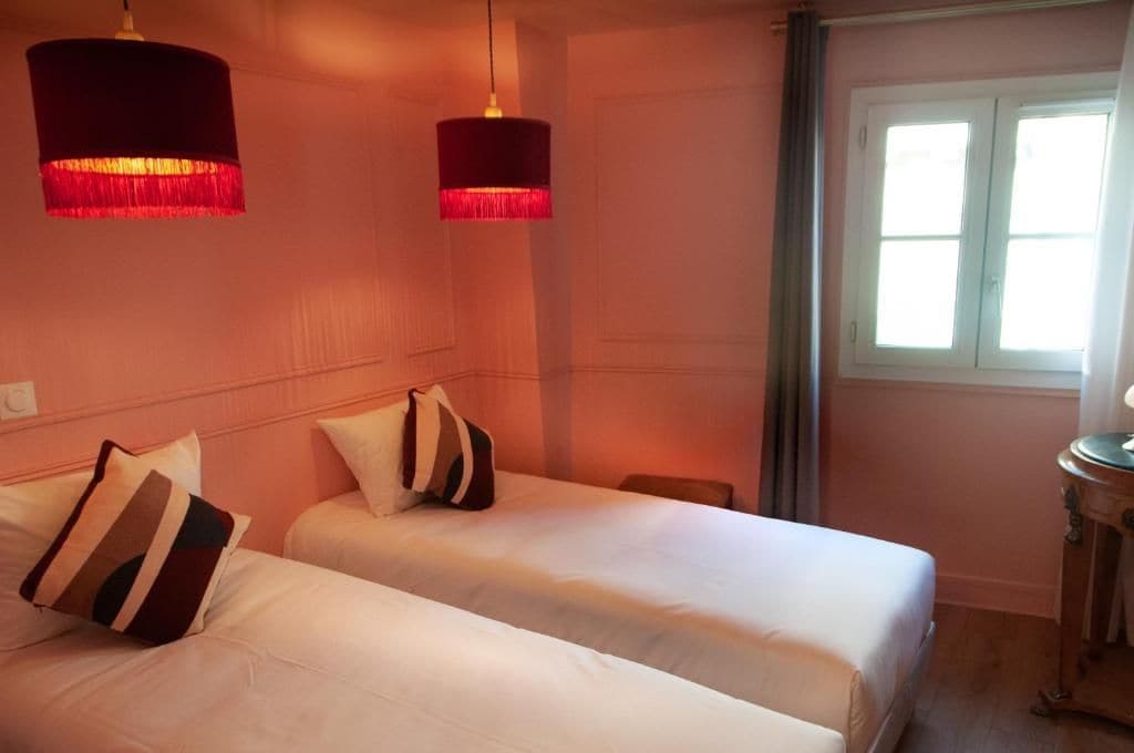 A hotel room with two beds, a window with curtains, and two pendant lights.